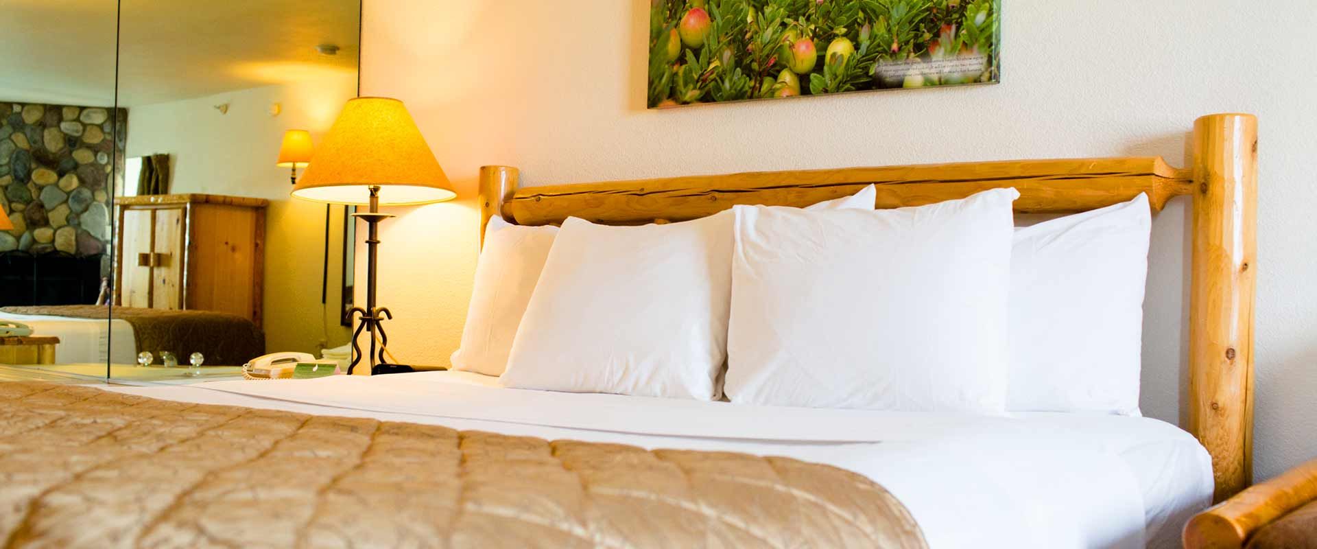 A hotel room with a bed, pillows, and a wooden headboard. A lit lamp sits on a nightstand.