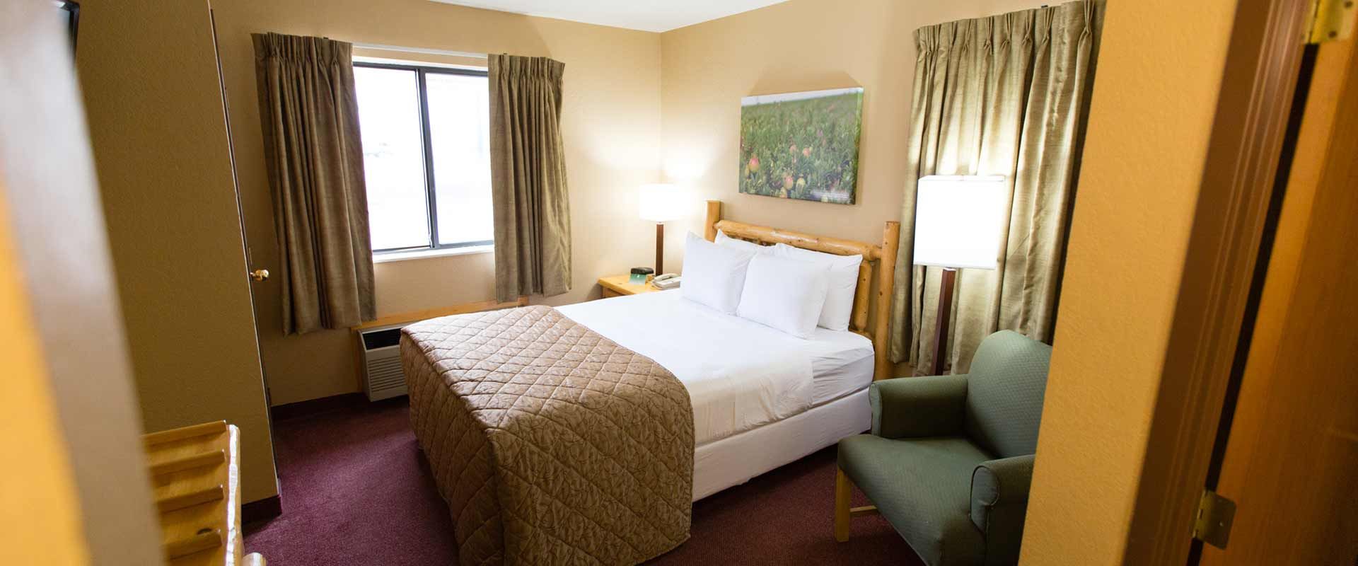 A hotel room with a bed, chair, window, and artwork. Brown, tan, and green colors are present.