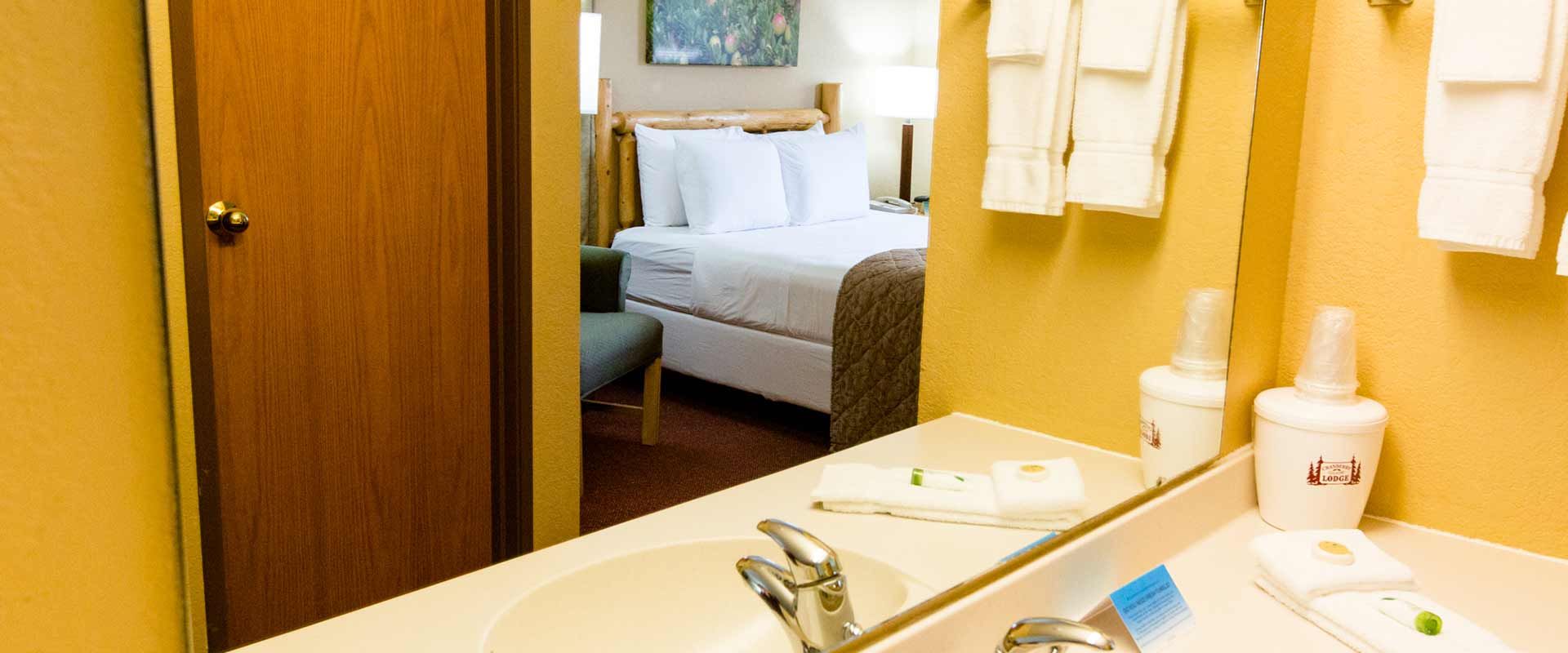 Bathroom with a sink and mirror reflecting a hotel bedroom with a bed and a partially open door.