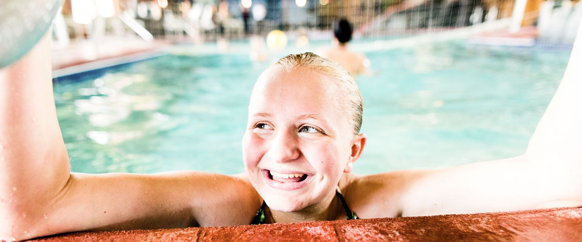 A person smiling in a swimming pool, resting their arms on the edge of the pool.