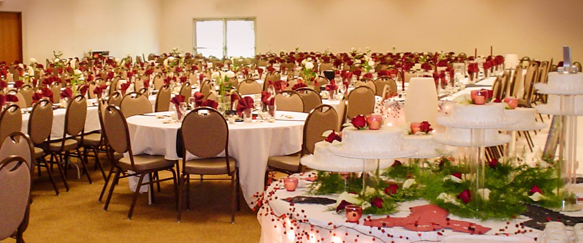 Wedding reception hall with round tables set for guests; cake display in the foreground.