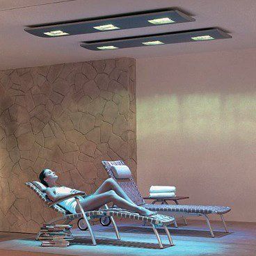 Frau entspannt unter Deckenleuchten in einem Spa-ähnlichen Raum mit Steinwand, Liegestühlen und Handtüchern.