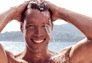 Lächelnder Mann, die Hände in den Haaren, mit strömendem Wasser, vor einem verschwommenen Wasserlandschaftshintergrund.