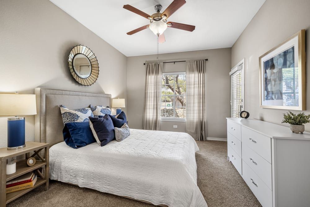 a bedroom with a bed, dresser, nightstand, mirror, and ceiling fan at Marquis at Deerfield in San Antonio, TX.