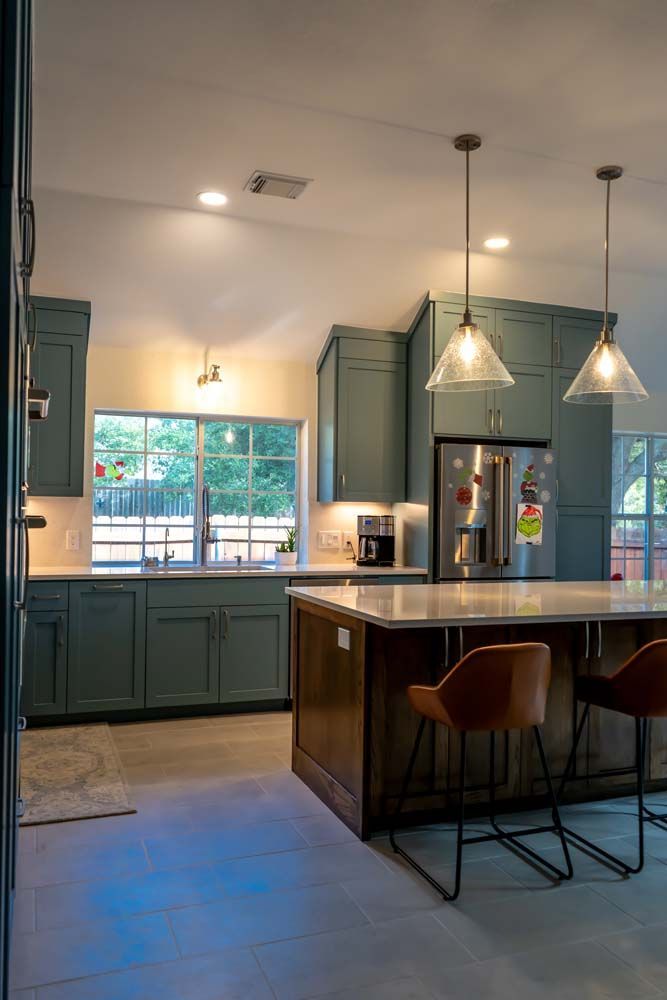 A kitchen with green cabinets , a large island , stools , a refrigerator , and a sink.