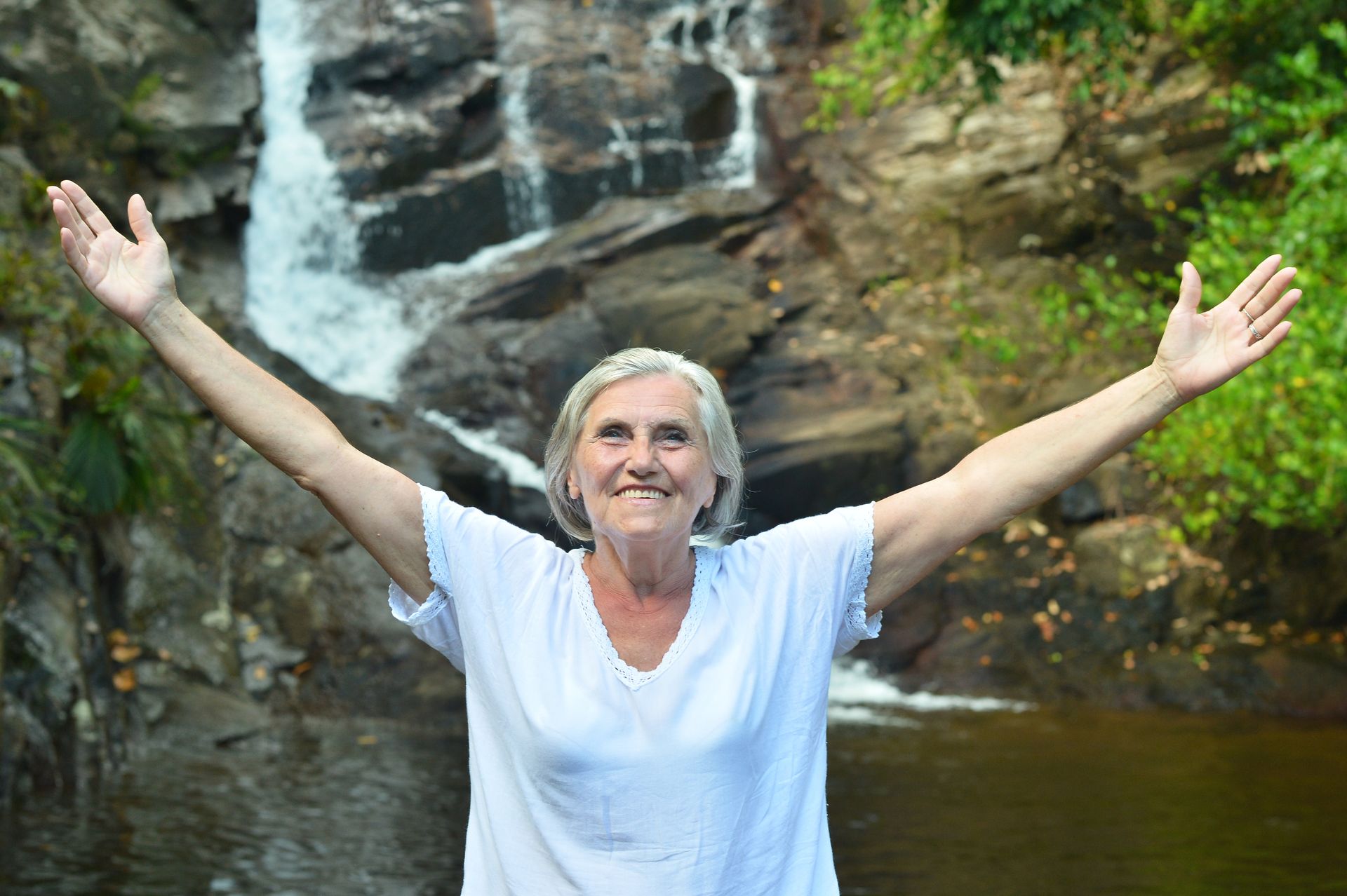 An elderly woman is standing in front of a waterfall with her arms outstretched.