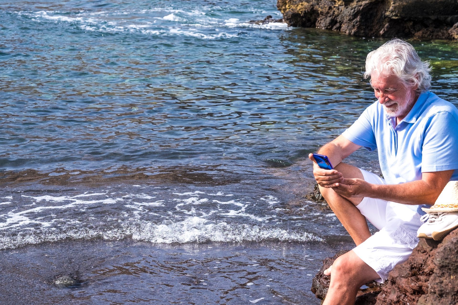 An older man is sitting on a rock near the ocean looking at his cell phone.