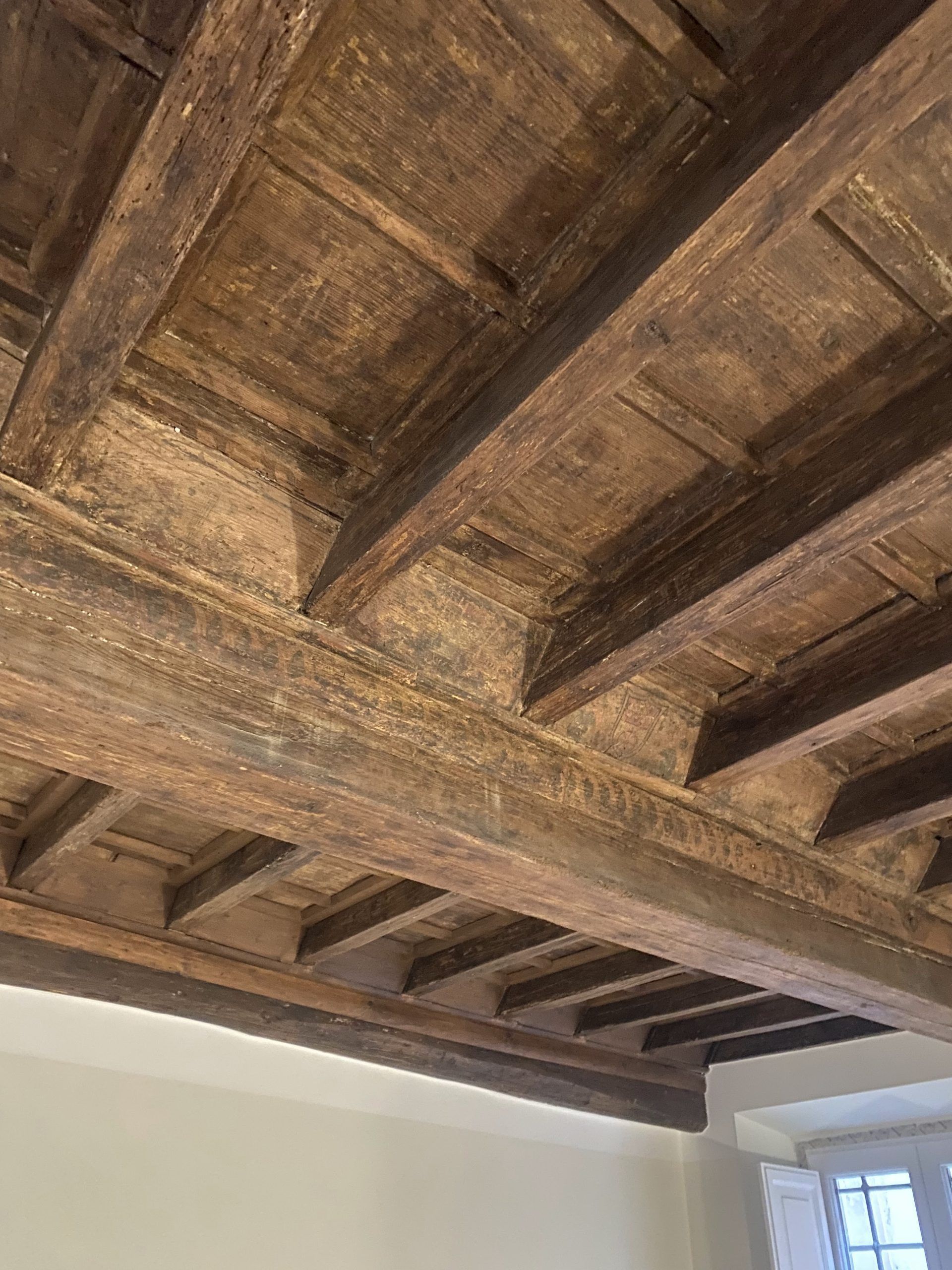 Primo piano di un soffitto rustico con travi in ​​legno, finestra e angolo di parete bianca sottostante.