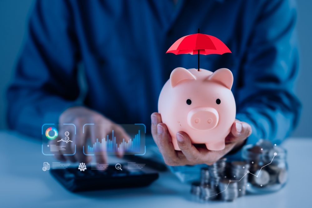 Person holding piggy bank under red umbrella; charts and coins visible, suggesting financial protection.