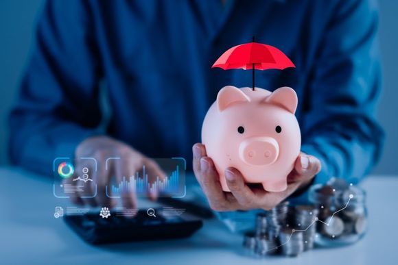 Person holding piggy bank under red umbrella; charts and coins visible, suggesting financial protection.