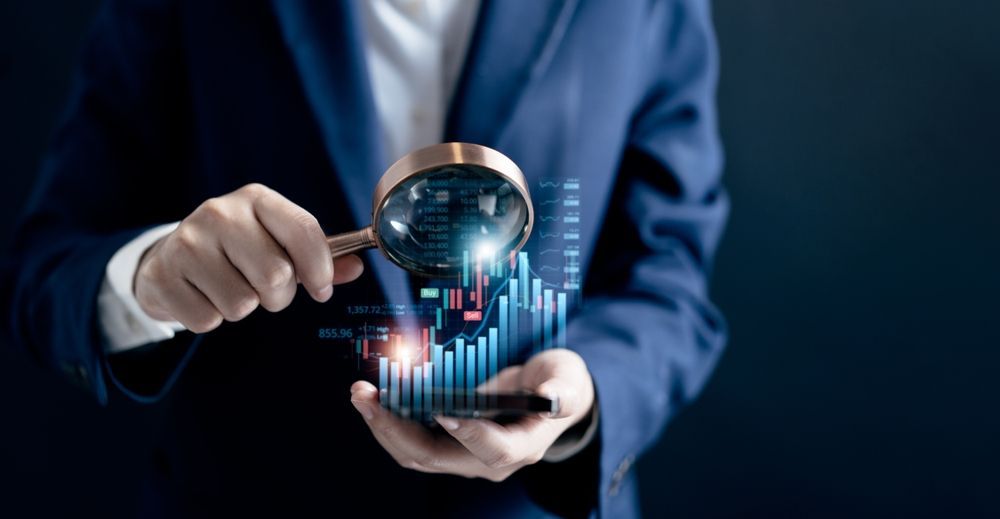 Person in suit holding magnifying glass over digital stock chart on a phone.