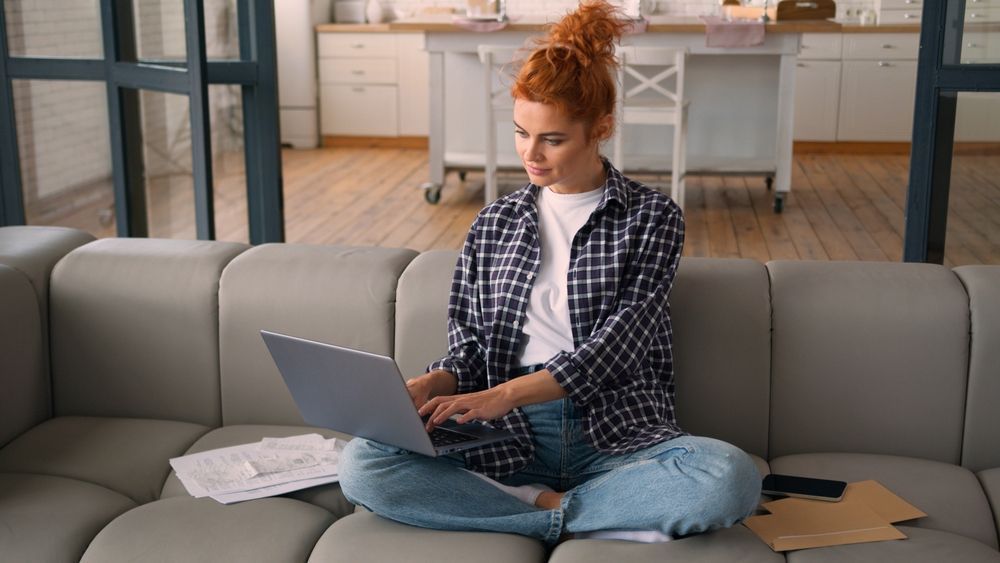 Woman with red hair works on laptop, sitting cross-legged on a gray couch with paperwork scattered.