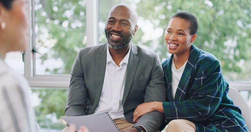 Couple smiling at a person holding papers indoors. They appear engaged, windows in background.
