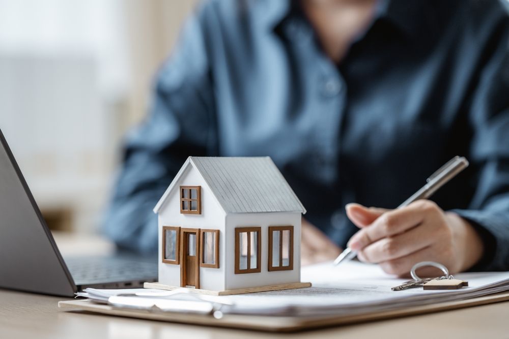 Person signing documents, small house model, laptop, and keys on a desk.