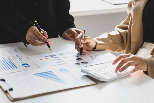 Two people analyze charts and graphs with pens and a calculator at a desk.
