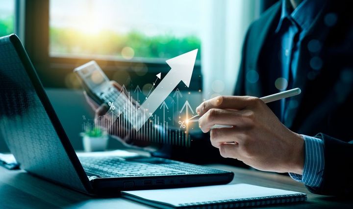 Businessperson using a laptop and calculator, analyzing data with a rising arrow graphic.