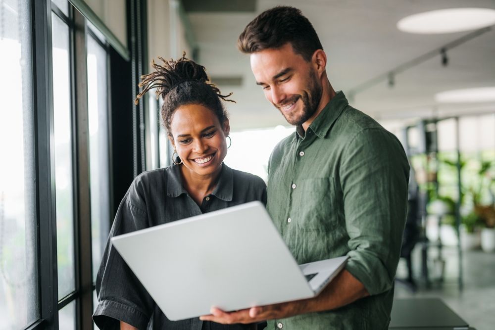 Two people looking at laptop, smiling near a window in an office.