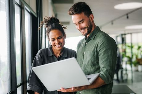 Two people looking at laptop, smiling near a window in an office.
