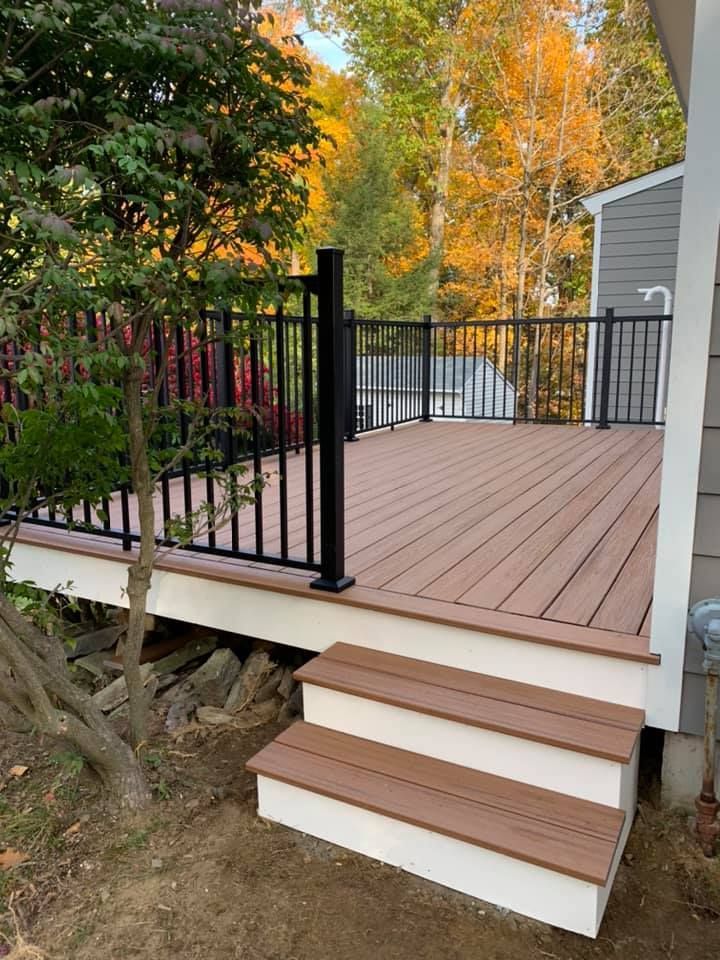 A brown composite deck with black metal railings and white trim, featuring two steps leading down to a dirt yard in autumn.