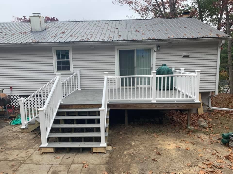 A grey-sided house with a metal roof features a grey wooden deck and stairs with white railings, leading to a patio area.