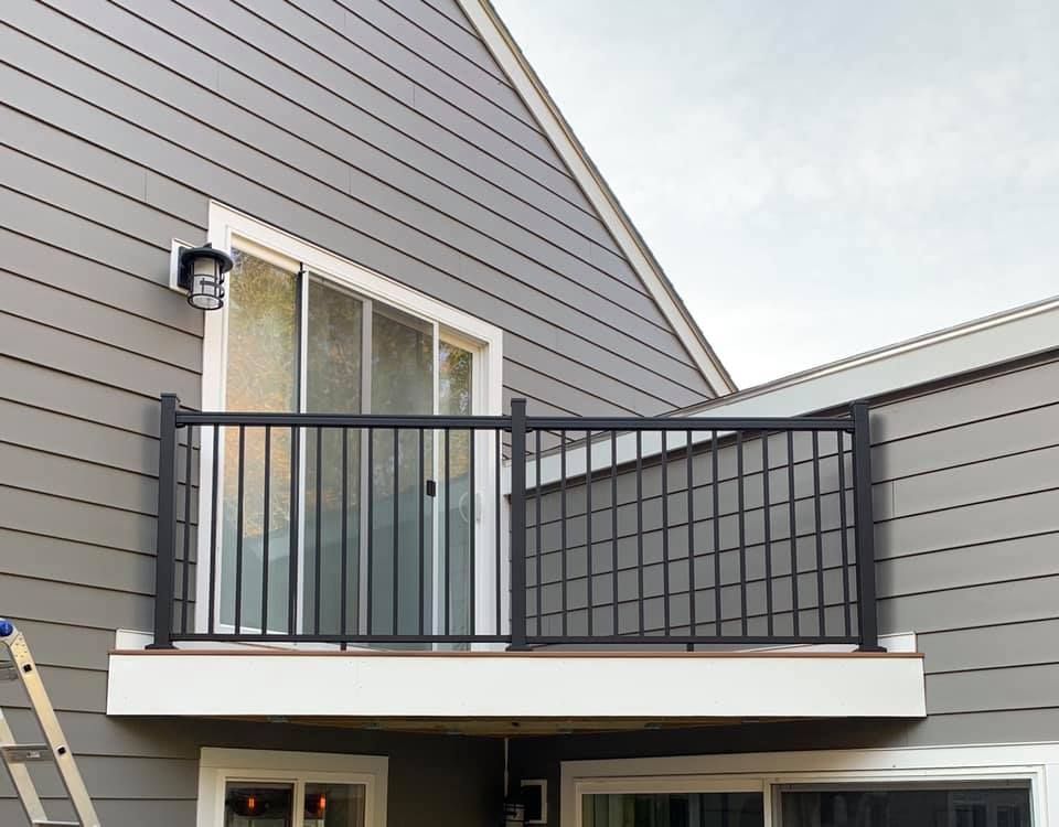 A balcony with a black metal railing attached to the side of a house with gray horizontal siding and a sliding glass door.