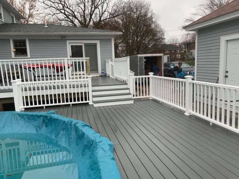 A gray composite deck surrounds an above-ground pool, featuring white railings and steps leading to a gray house exterior.