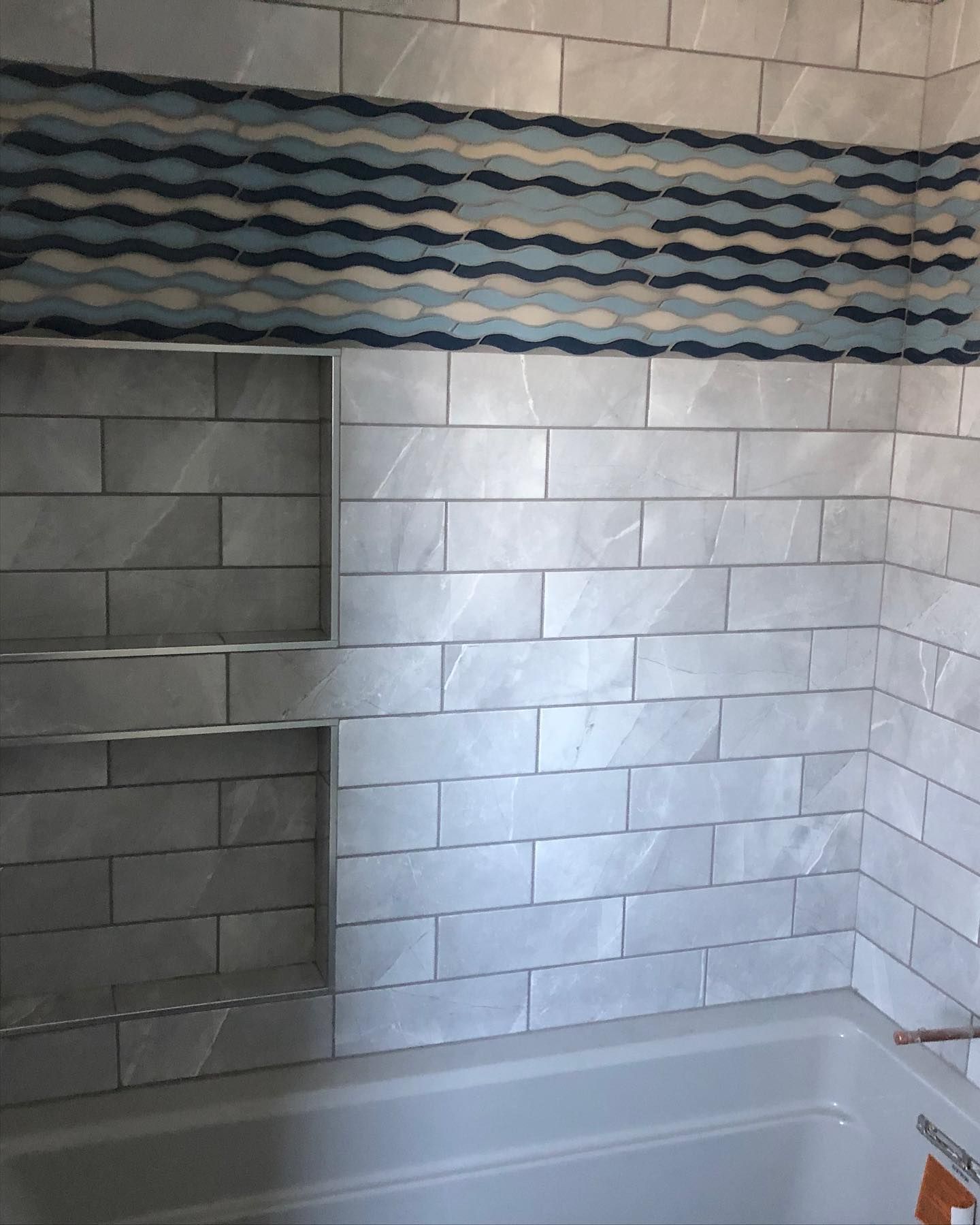A bathroom shower wall with white rectangular tiles, two built-in recessed niches, and a blue decorative wave-pattern band.