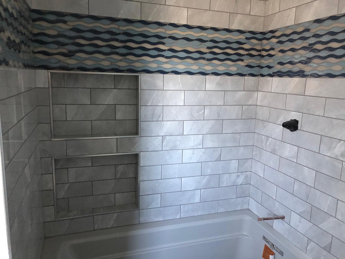 A shower wall with white rectangular tiles, a dual-niche storage area, and a decorative blue wavy-patterned accent stripe.