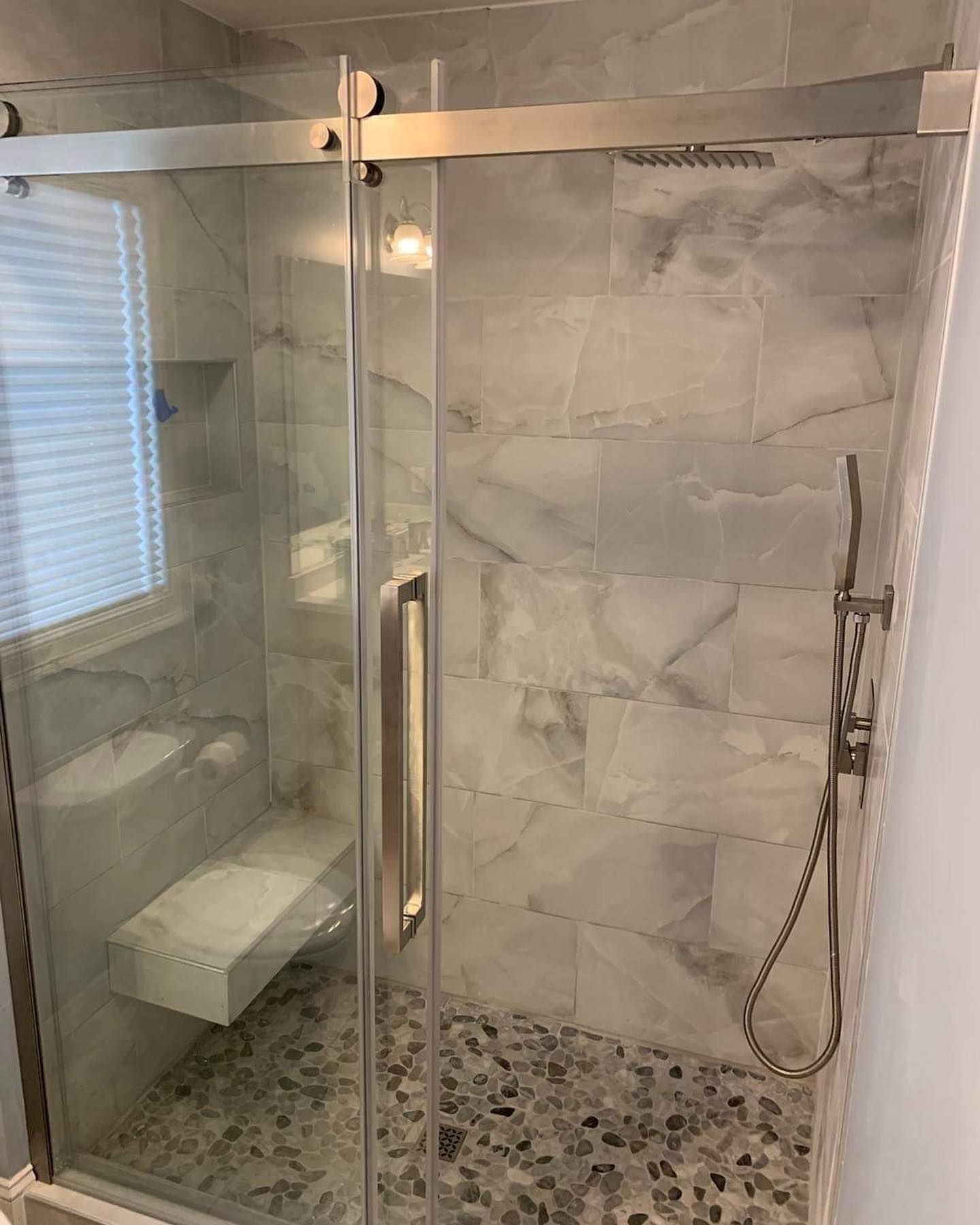 A modern walk-in shower with a sliding glass door, white marble wall tiles, a pebble floor, and a handheld shower head.