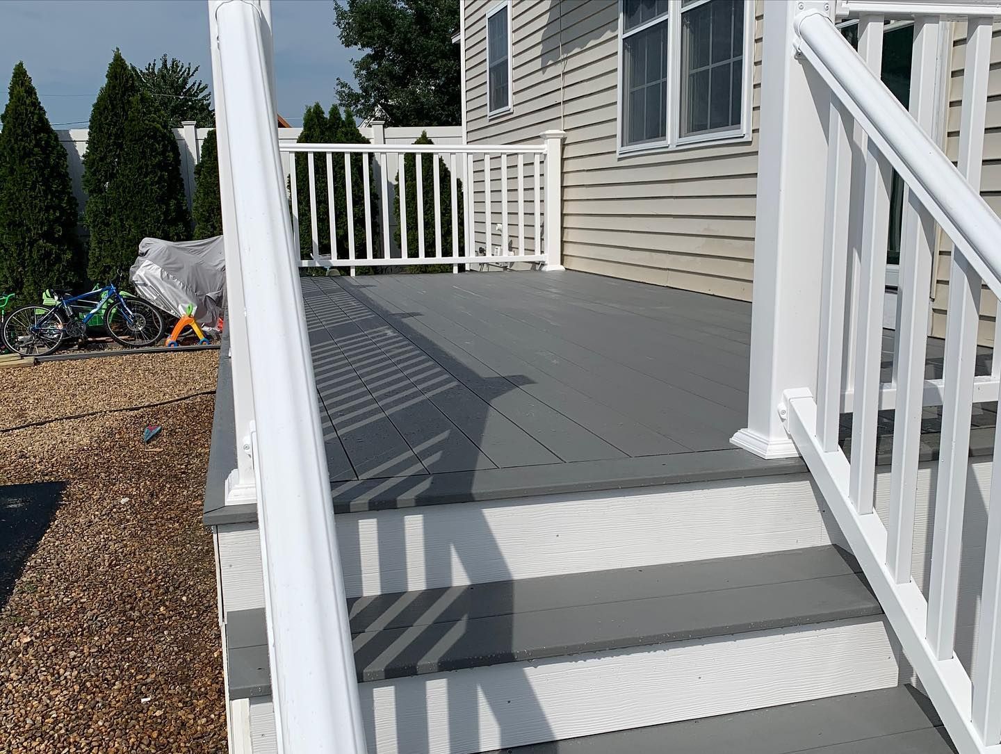 A gray-decked porch with white railings and stairs leads to the side entrance of a beige, vinyl-sided home.