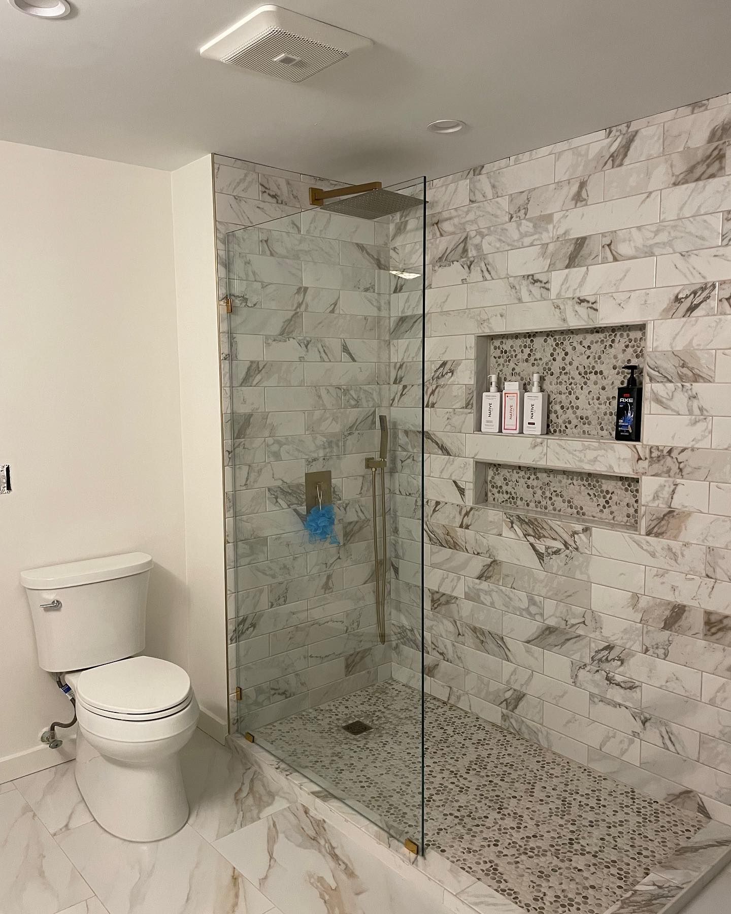A modern bathroom with a glass-enclosed shower featuring marble-look tile, pebble flooring, and built-in shelving.