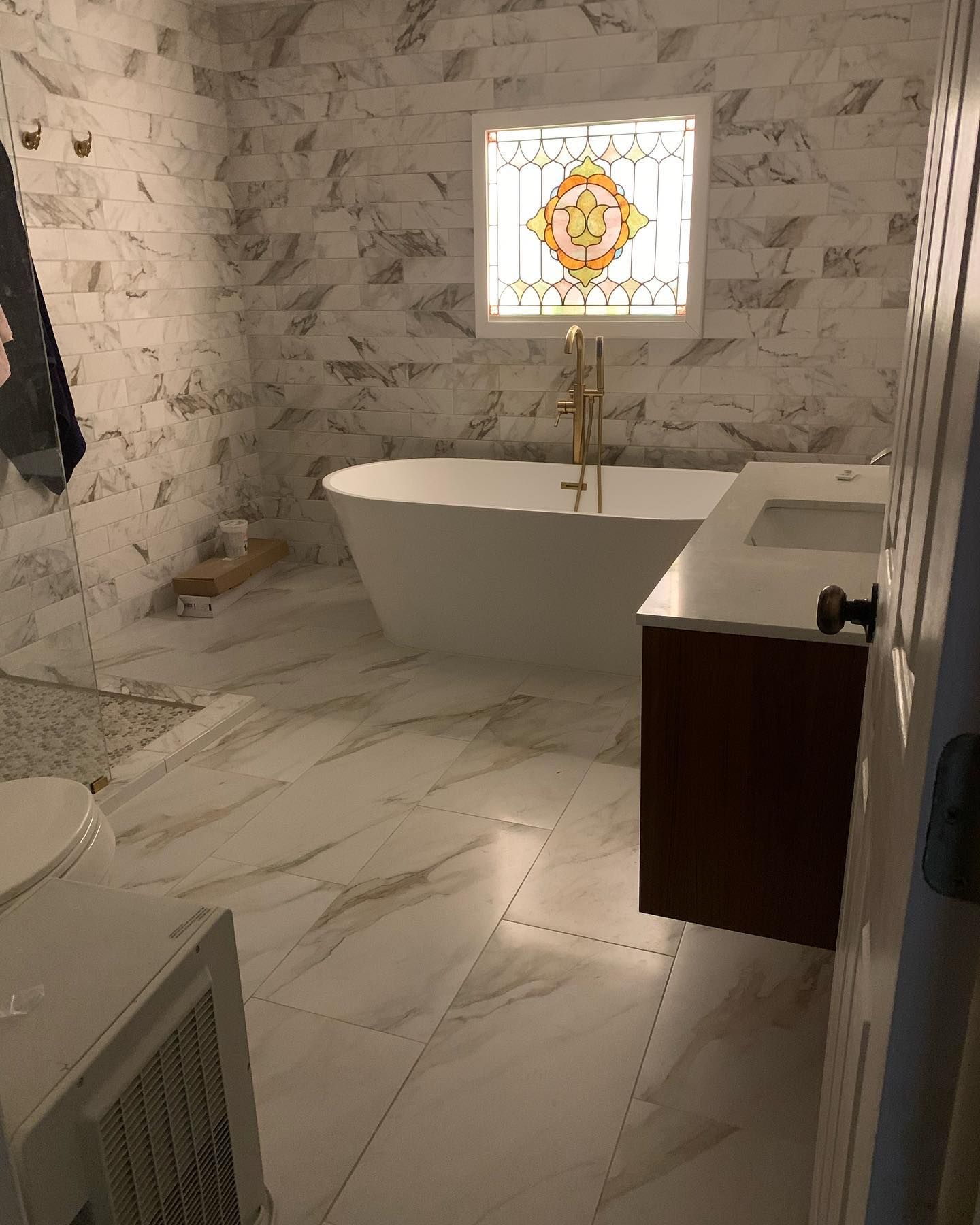 A bright bathroom with white marble tile walls and floors, a freestanding white tub, and a stained glass window.