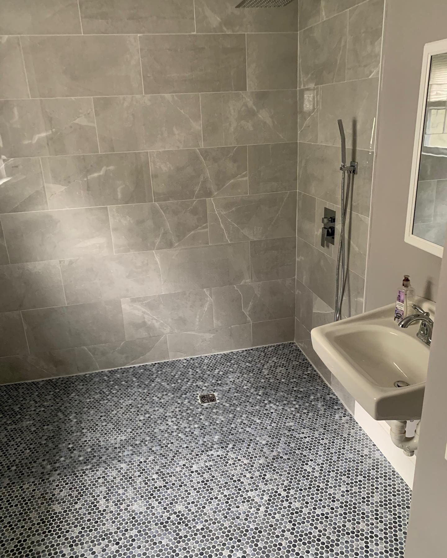 A shower area with grey stone-look wall tiles, a dark pebble-texture floor, a wall-mounted sink, and a handheld shower head.