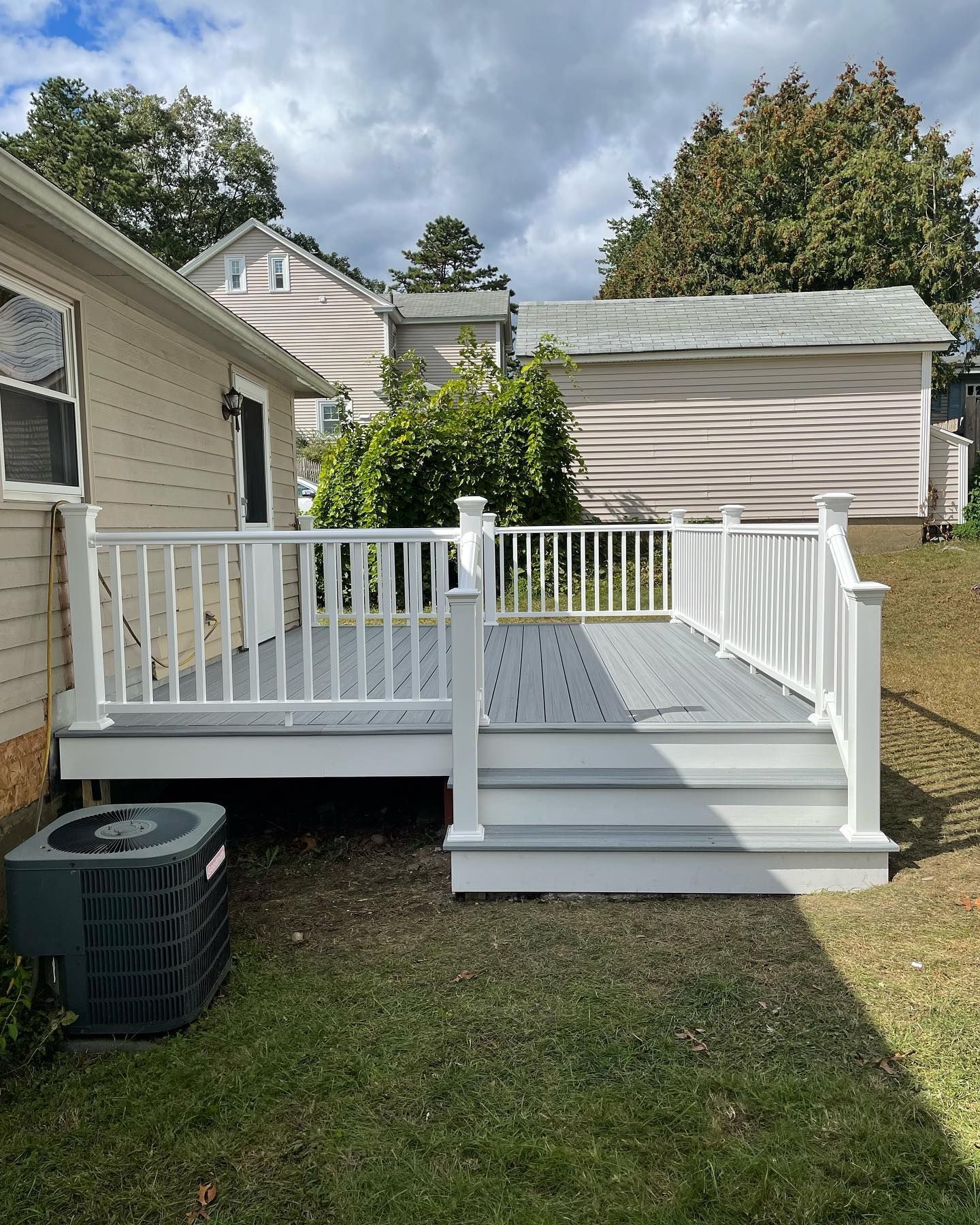 A gray backyard deck with white railings and stairs attached to a beige house next to a central air conditioning unit.