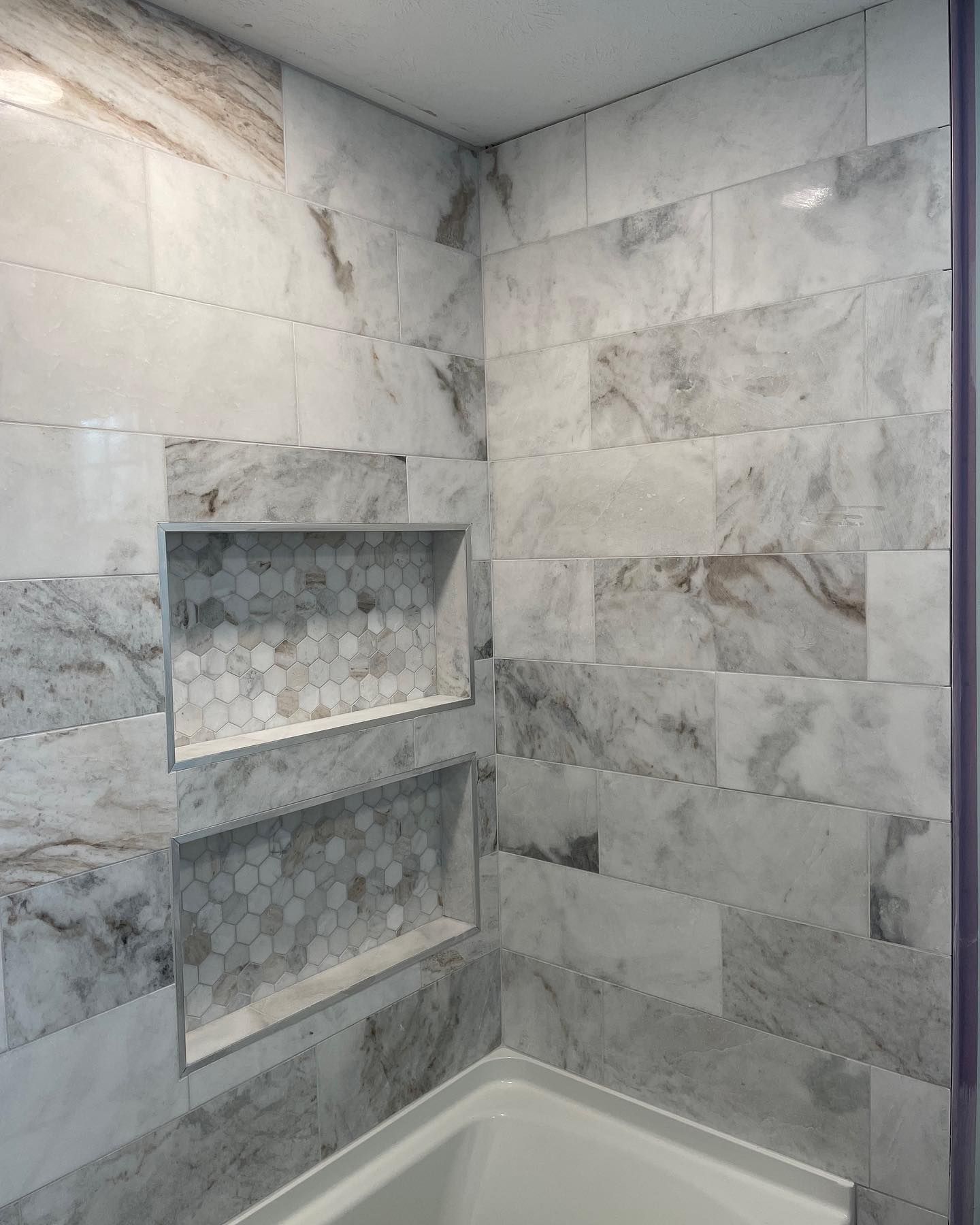 A modern bathroom shower wall tiled in gray and white marble with two built-in recessed niches featuring hexagonal tiles.