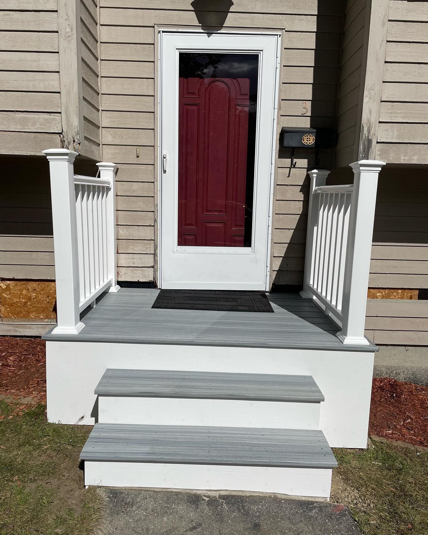 A red front door with a storm door, flanked by white railings, leads to a gray-painted concrete porch with two steps.