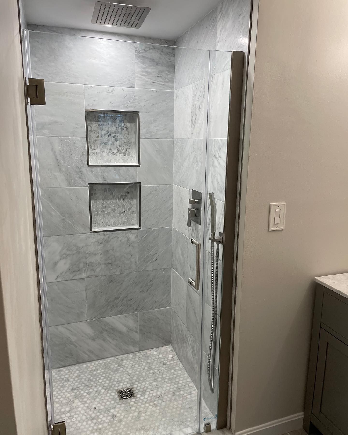 A walk-in shower with gray marbled tile walls, a pebbled floor, dual wall niches, and a glass door with metal hardware.