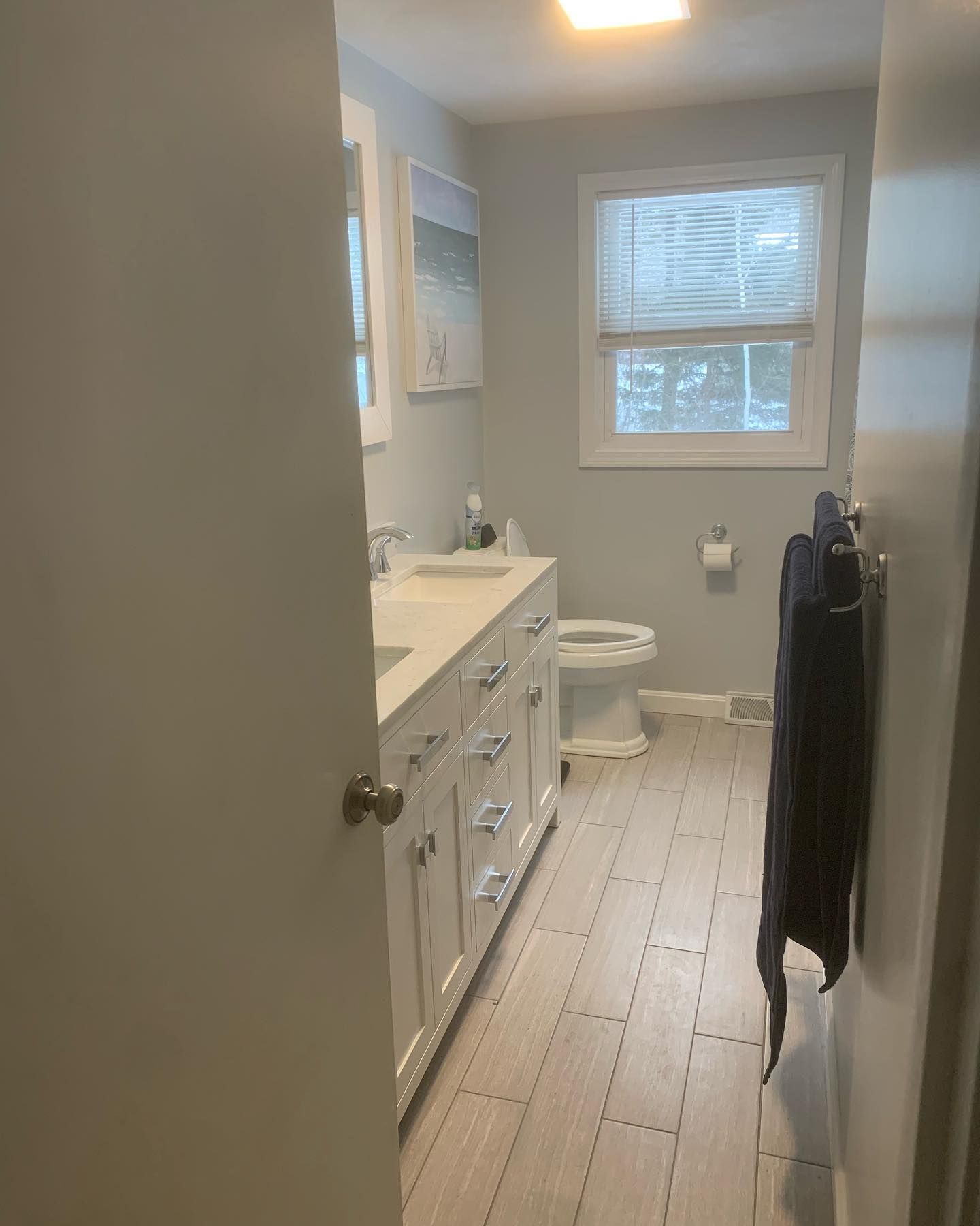 A bathroom with a white vanity, a toilet, a window with blinds, light gray walls, and light-colored wood-look floor tiles.