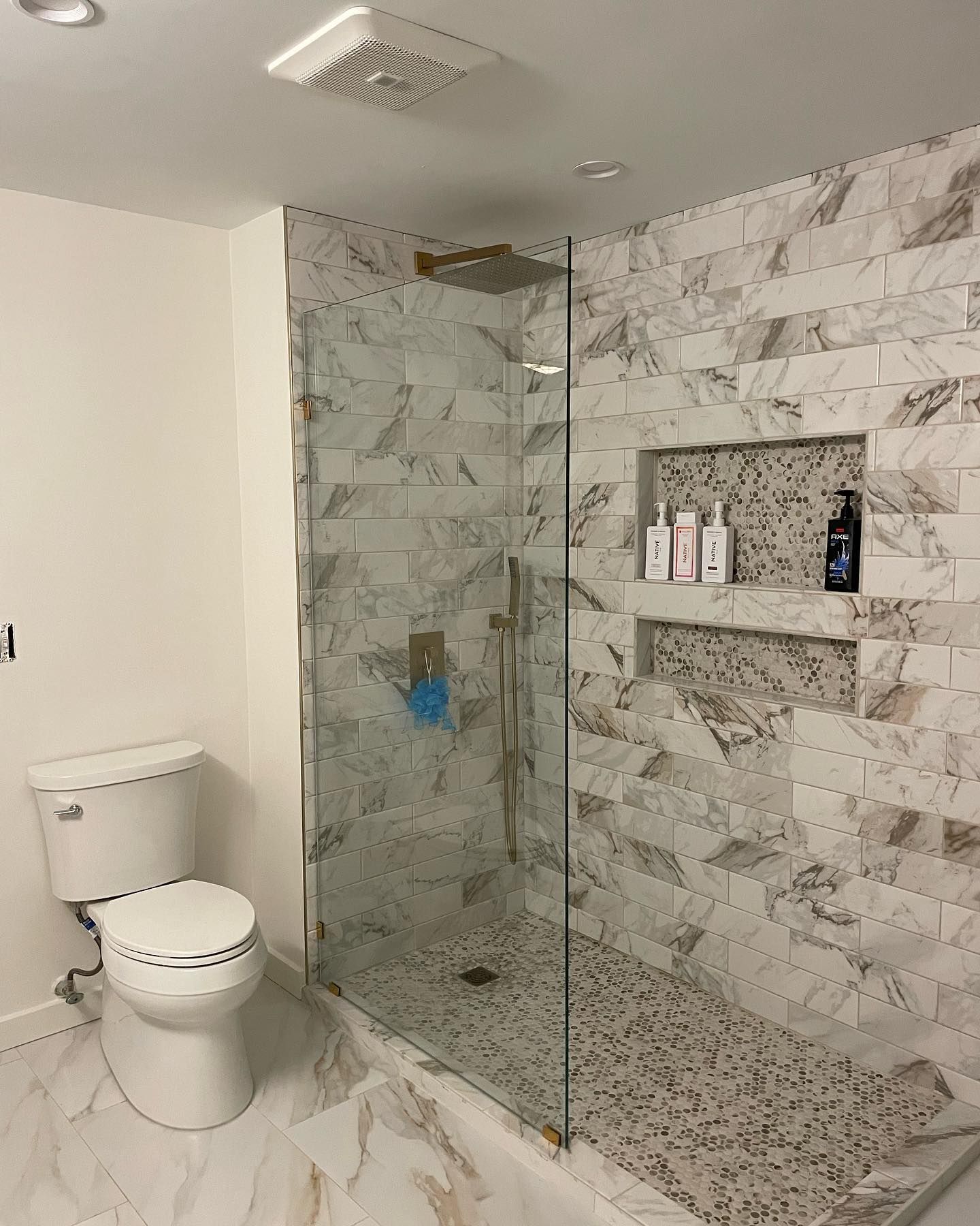 Modern bathroom with a white toilet next to a glass-enclosed shower featuring marble wall tile and pebble flooring.
