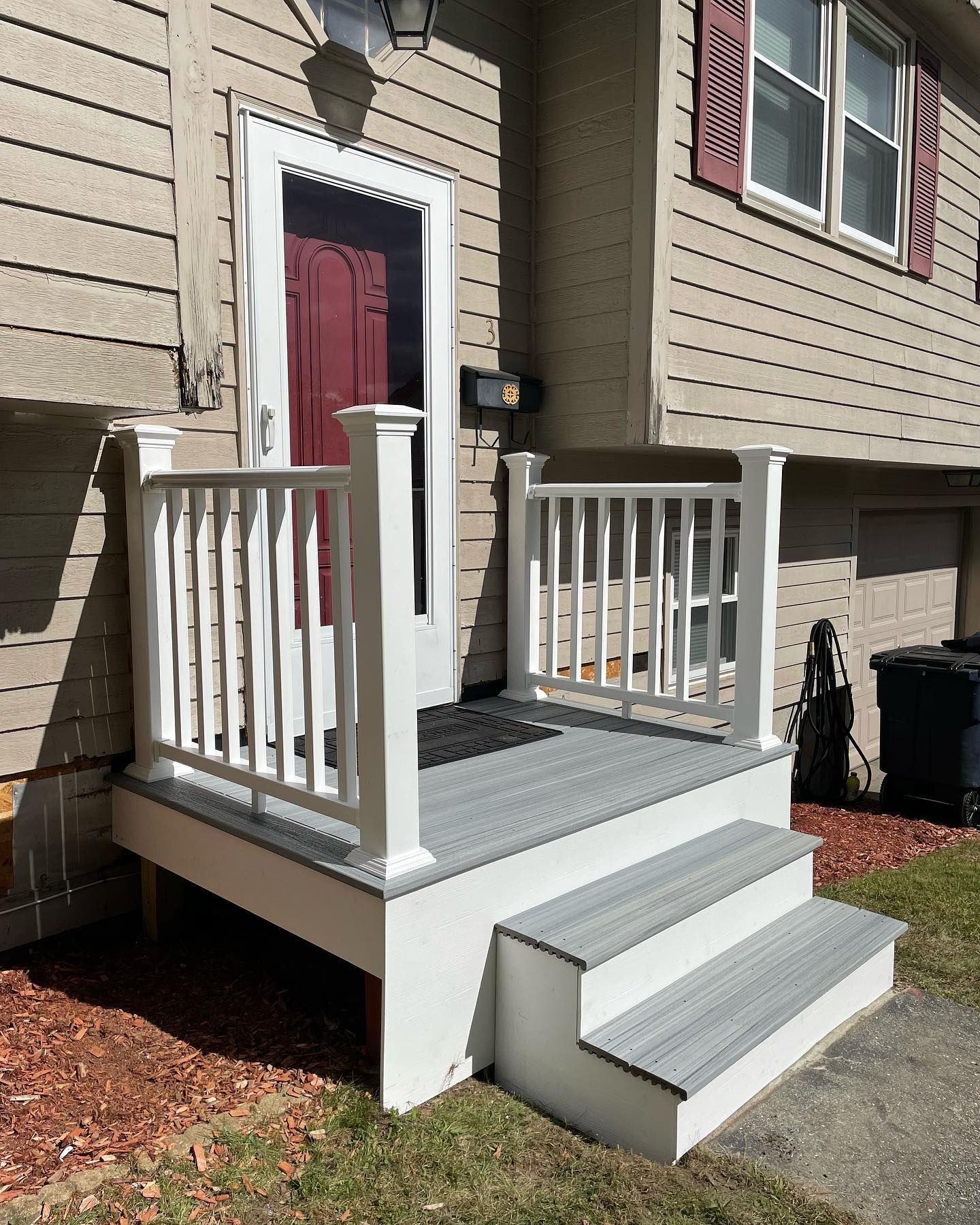 A small, elevated deck with light gray composite decking and white railings leading to a red front door of a beige house.