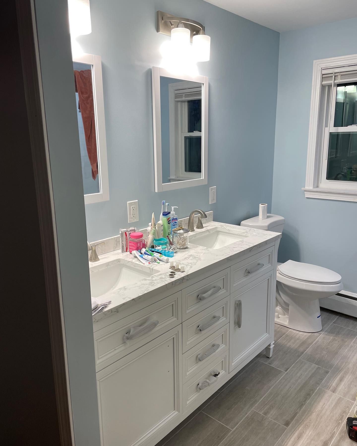 A bathroom with a white double vanity, light blue walls, two mirrors, a light fixture, and a toilet on tiled flooring.