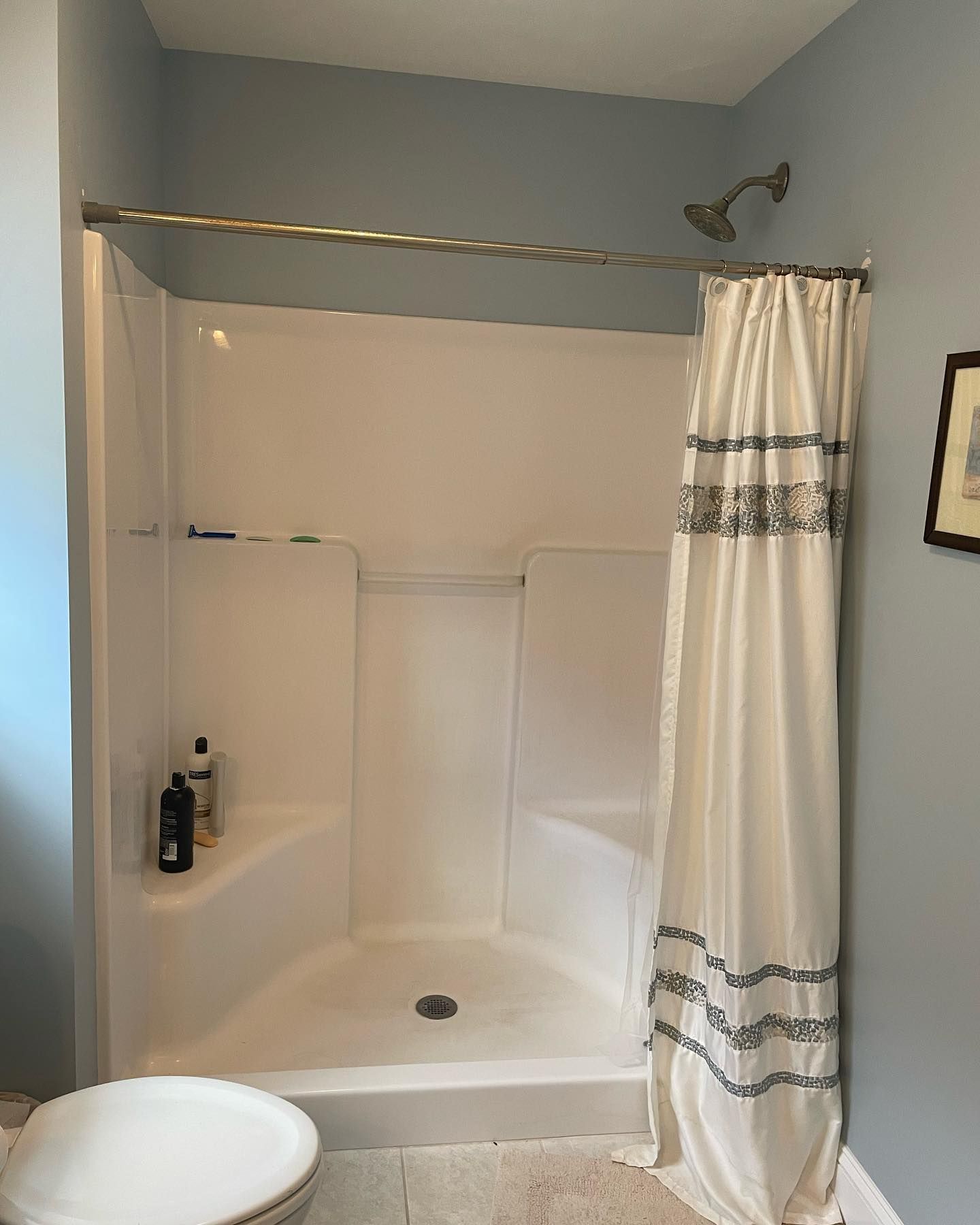 A white walk-in shower stall with built-in corner seats, a metal curtain rod, and a white curtain with grey trim.