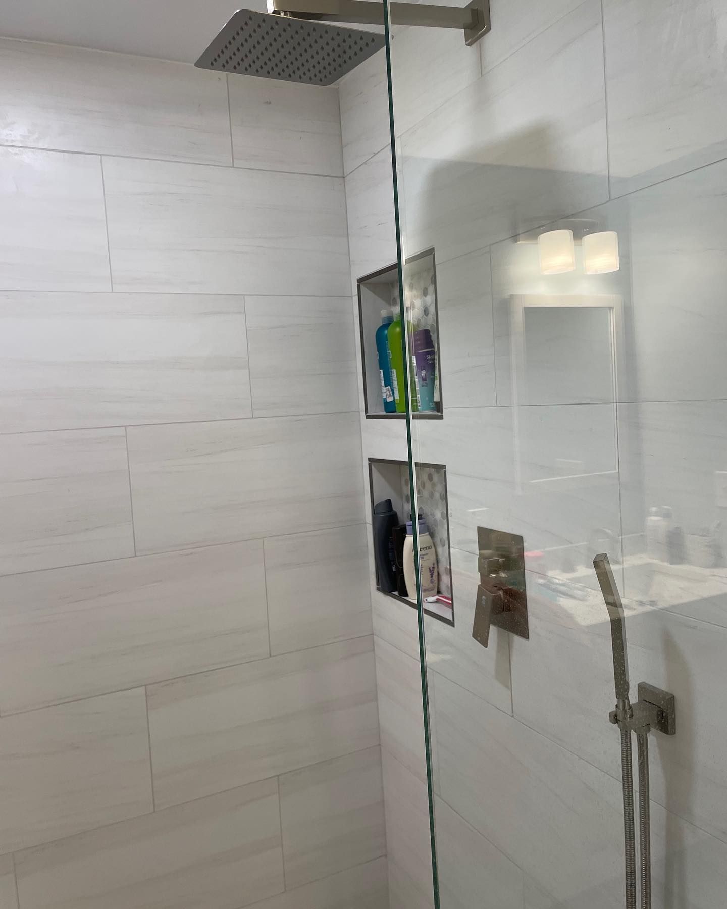 A modern glass-enclosed shower with white tiled walls, a rain showerhead, built-in niches, and brushed metal fixtures.