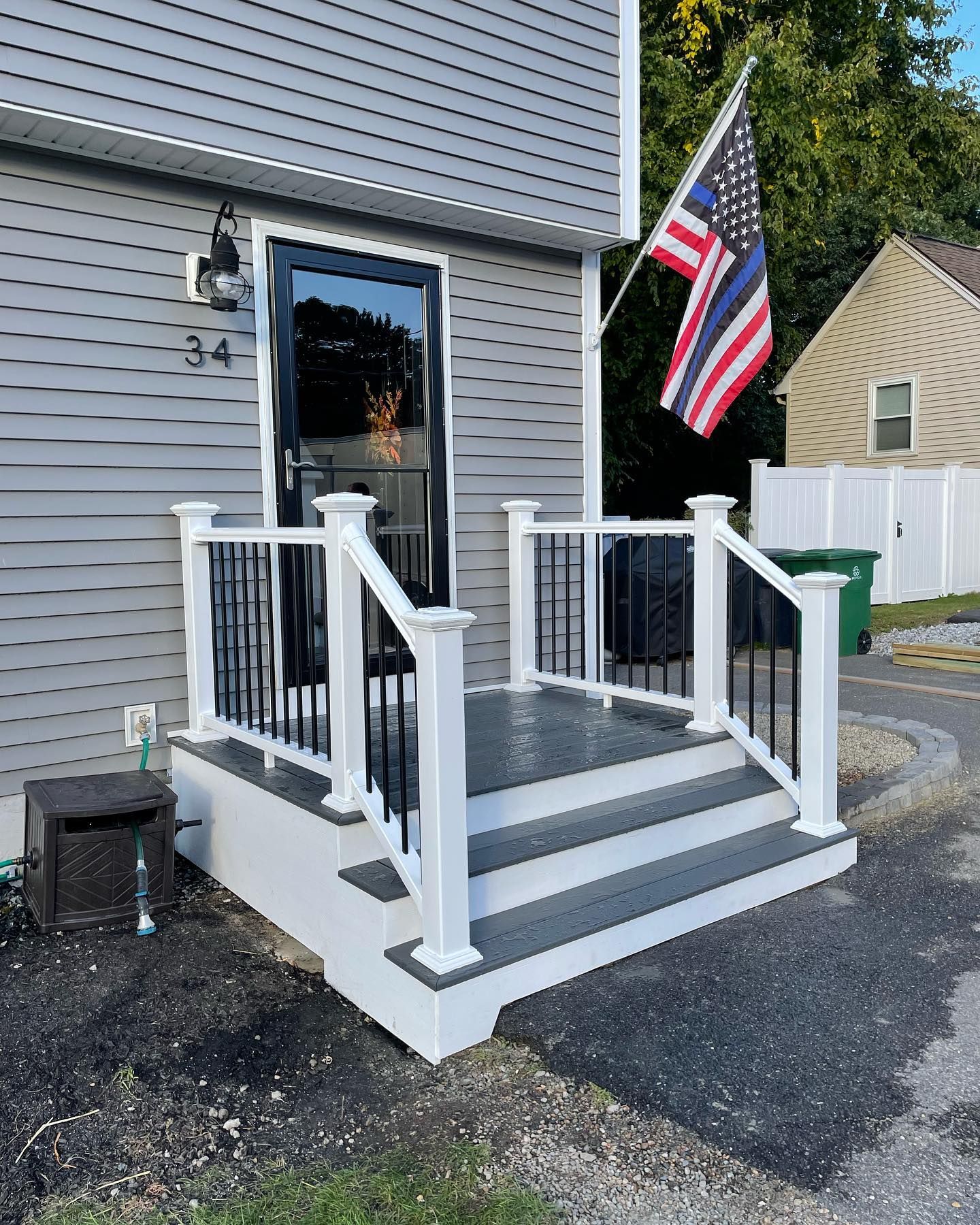 A grey house with a white front porch, black steps, railings, and a thin blue line American flag hanging by the door.