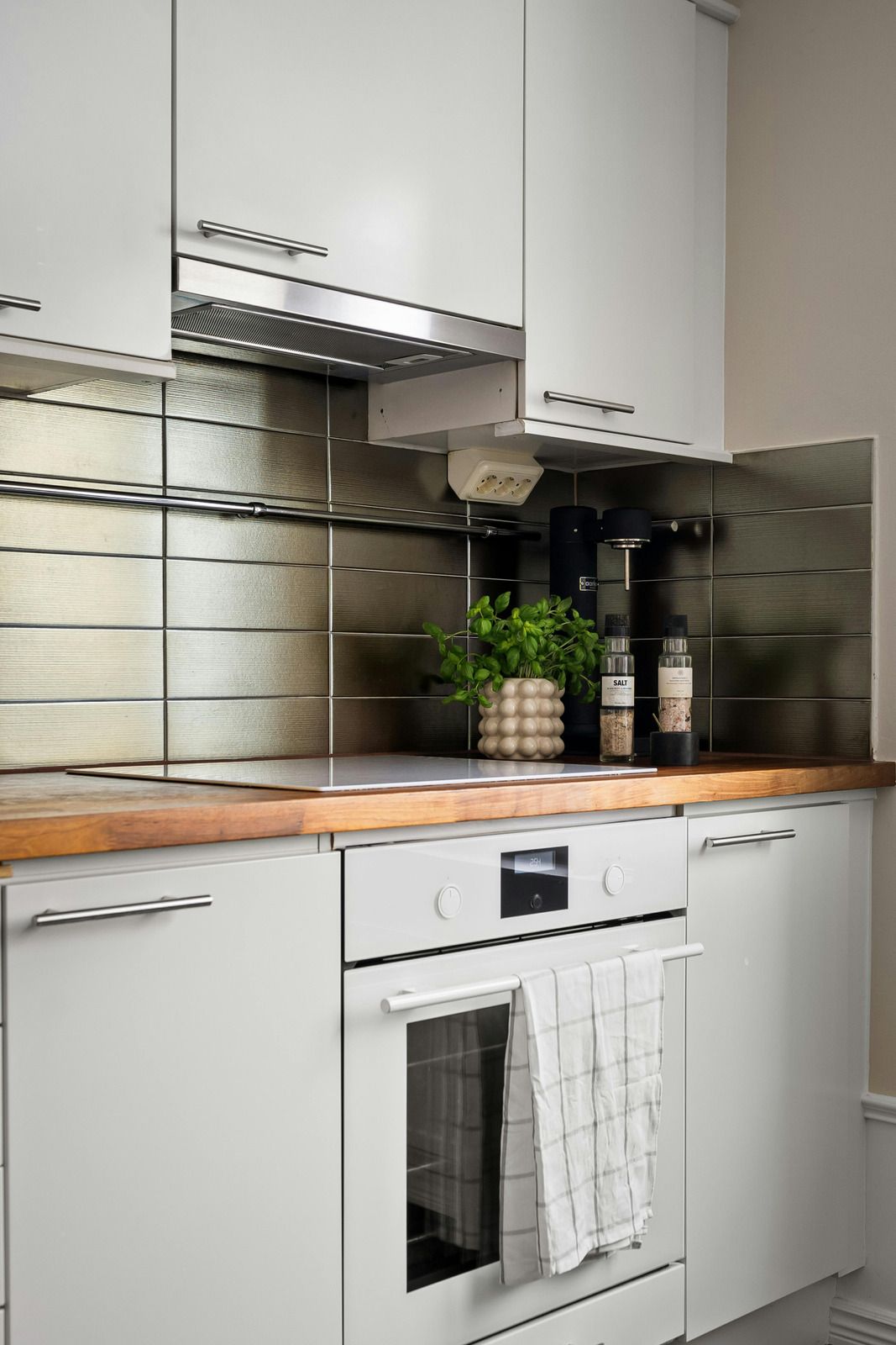 A white kitchen cabinet and oven setup with a wooden countertop and metallic horizontal-tiled backsplash.