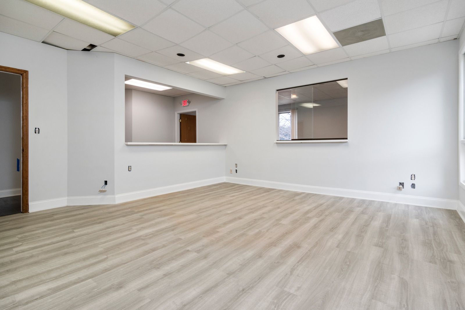 Empty office room with light grey walls, wood-look flooring, two interior windows, and a doorway on the left.
