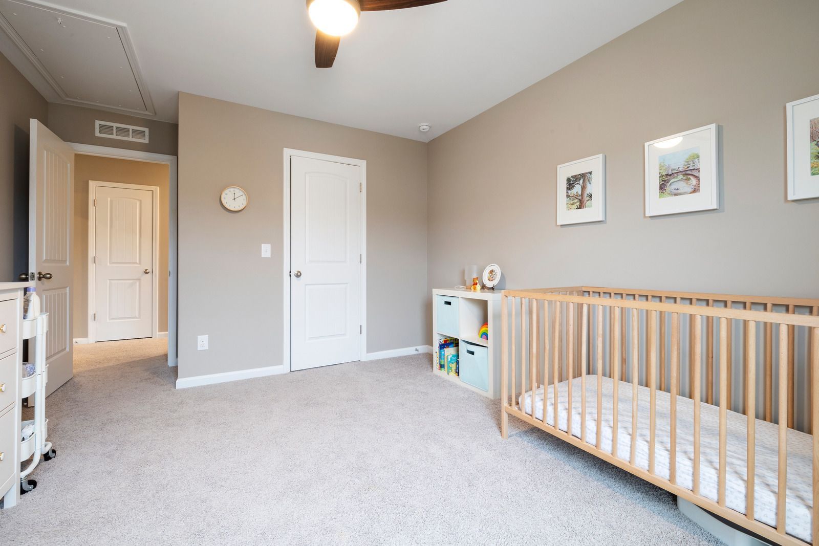 A nursery with beige walls, light carpet, a wooden crib, a toy organizer, and two doors leading to other rooms.