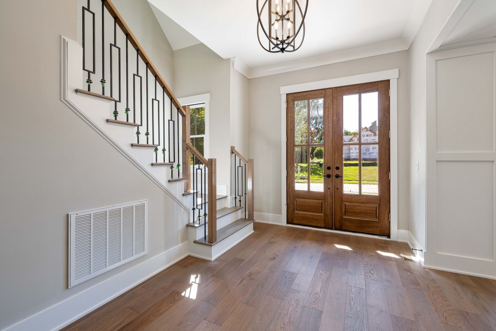 Bright entryway featuring wood floors, a staircase with decorative metal railing, a white vent, and wooden double doors.