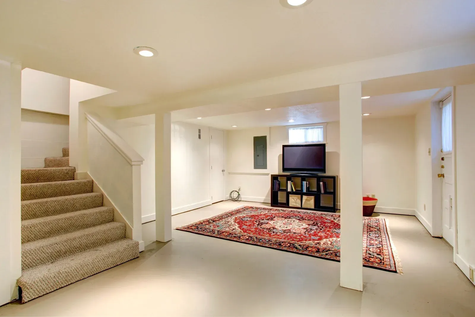 A finished basement with beige carpeted stairs, a central pillar, a television on a stand, and a red patterned area rug.