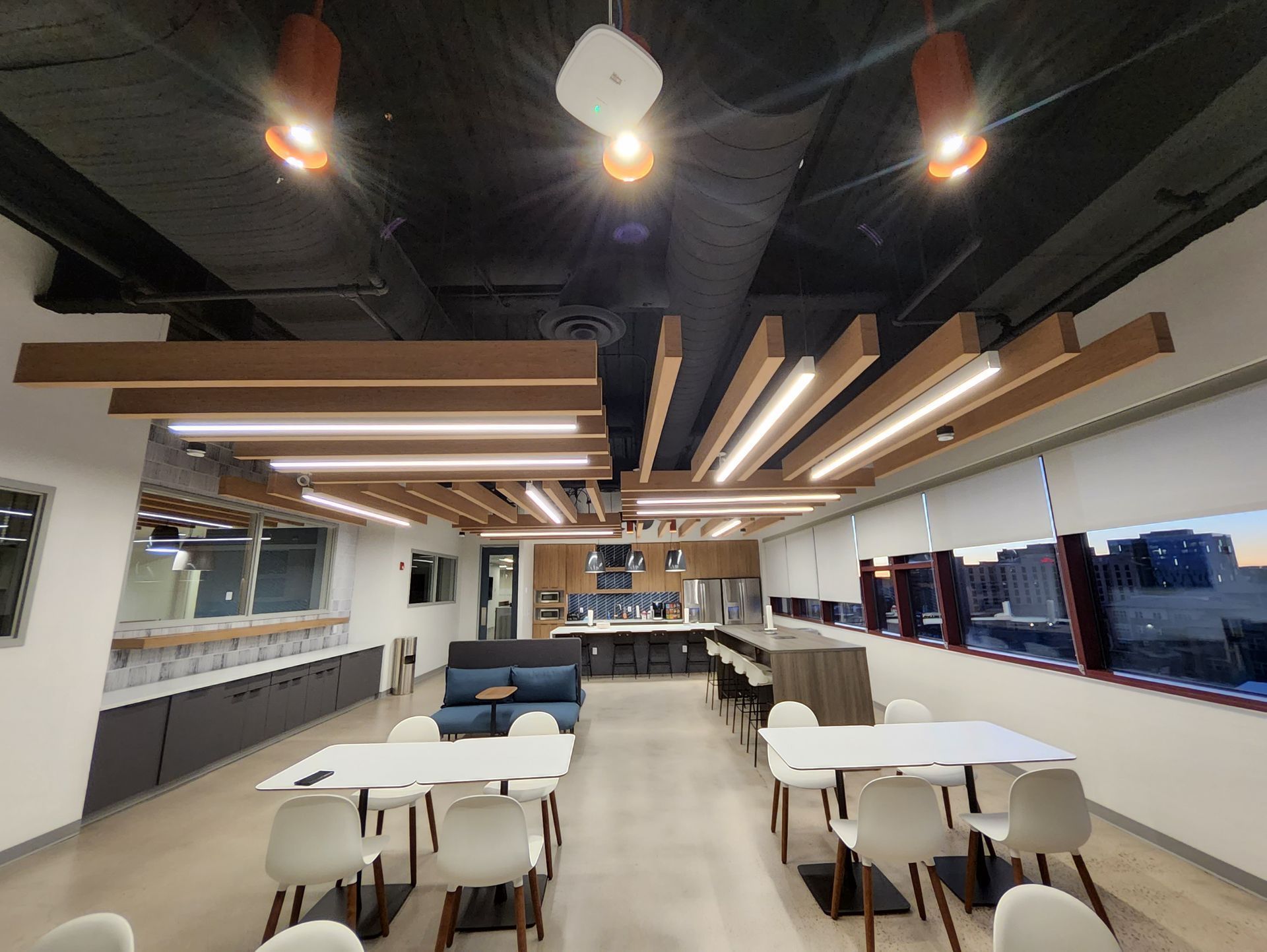 Modern office kitchen and dining area with tables, chairs, and decorative ceiling lighting.